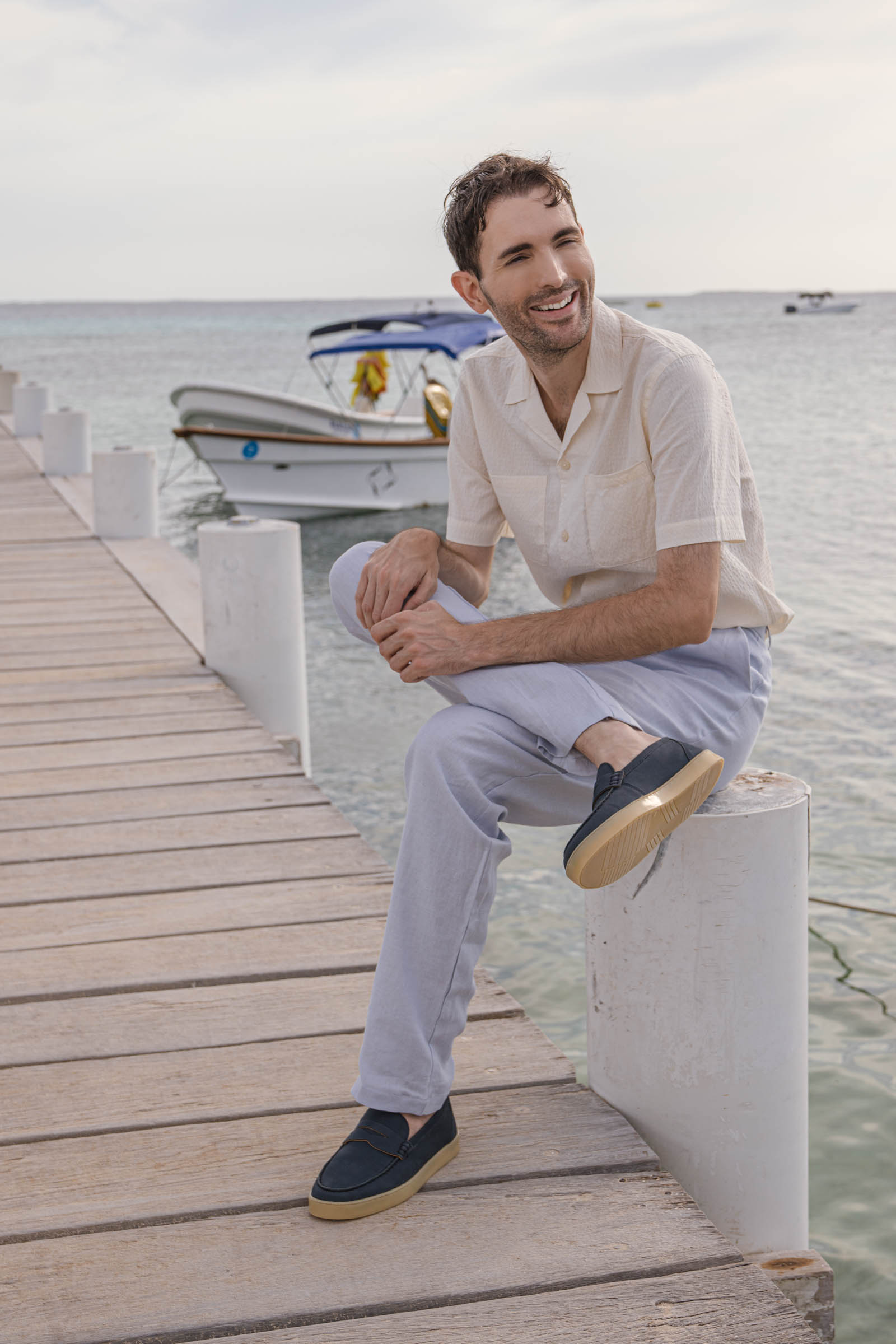 Marko Tadic blogger in complete Jil Sander look with beige shirt, light blue linen pants and blue suede loafers demonstrates timeless fashion on wooden pier surrounded by crystal-clear Caribbean sea waters