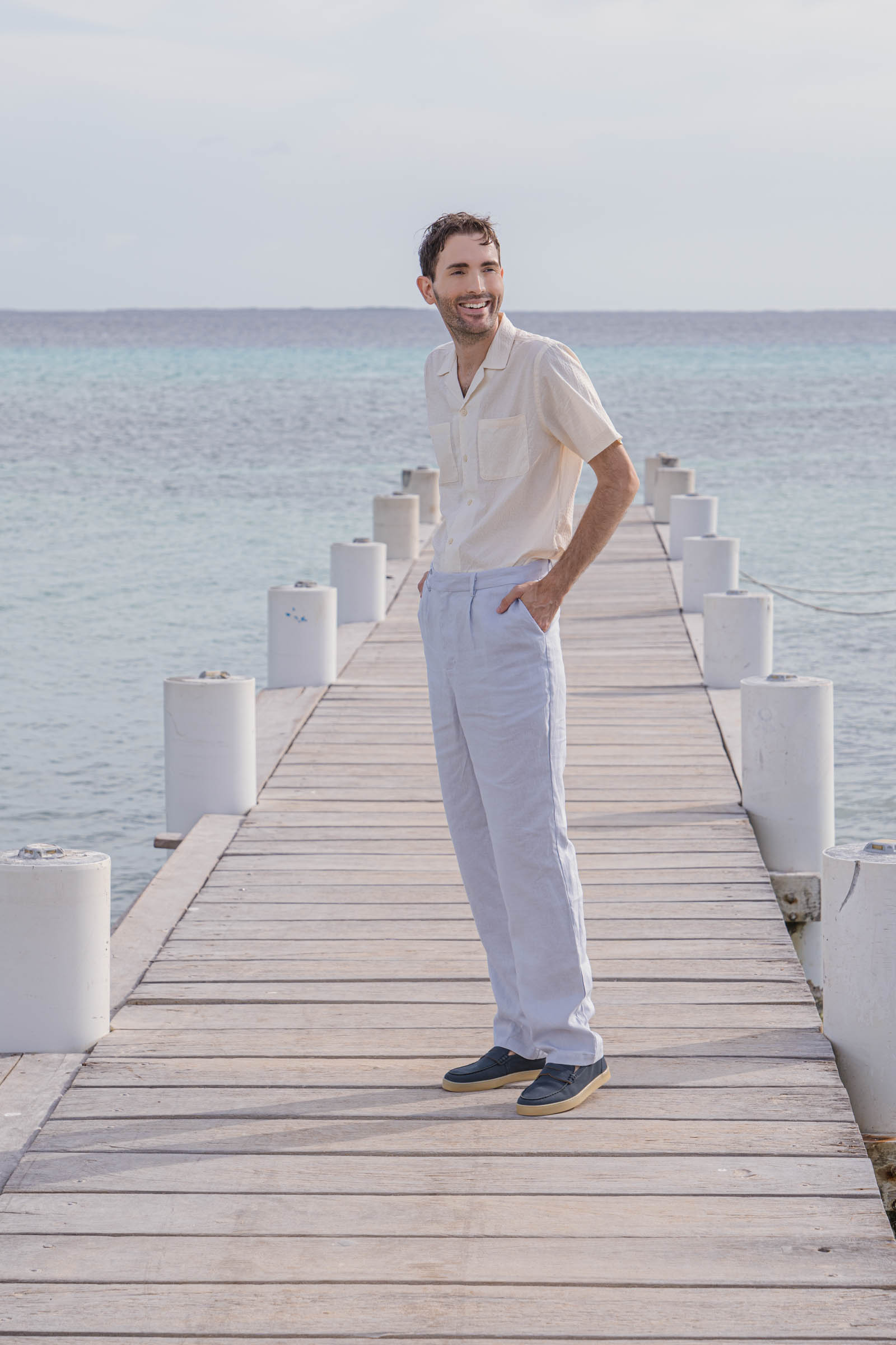 Marko Tadic owner of Mr.M by Marko Tadic blog poses on wooden dock in Los Roques archipelago wearing complete Jil Sander outfit with beige short-sleeve shirt and light blue linen pants