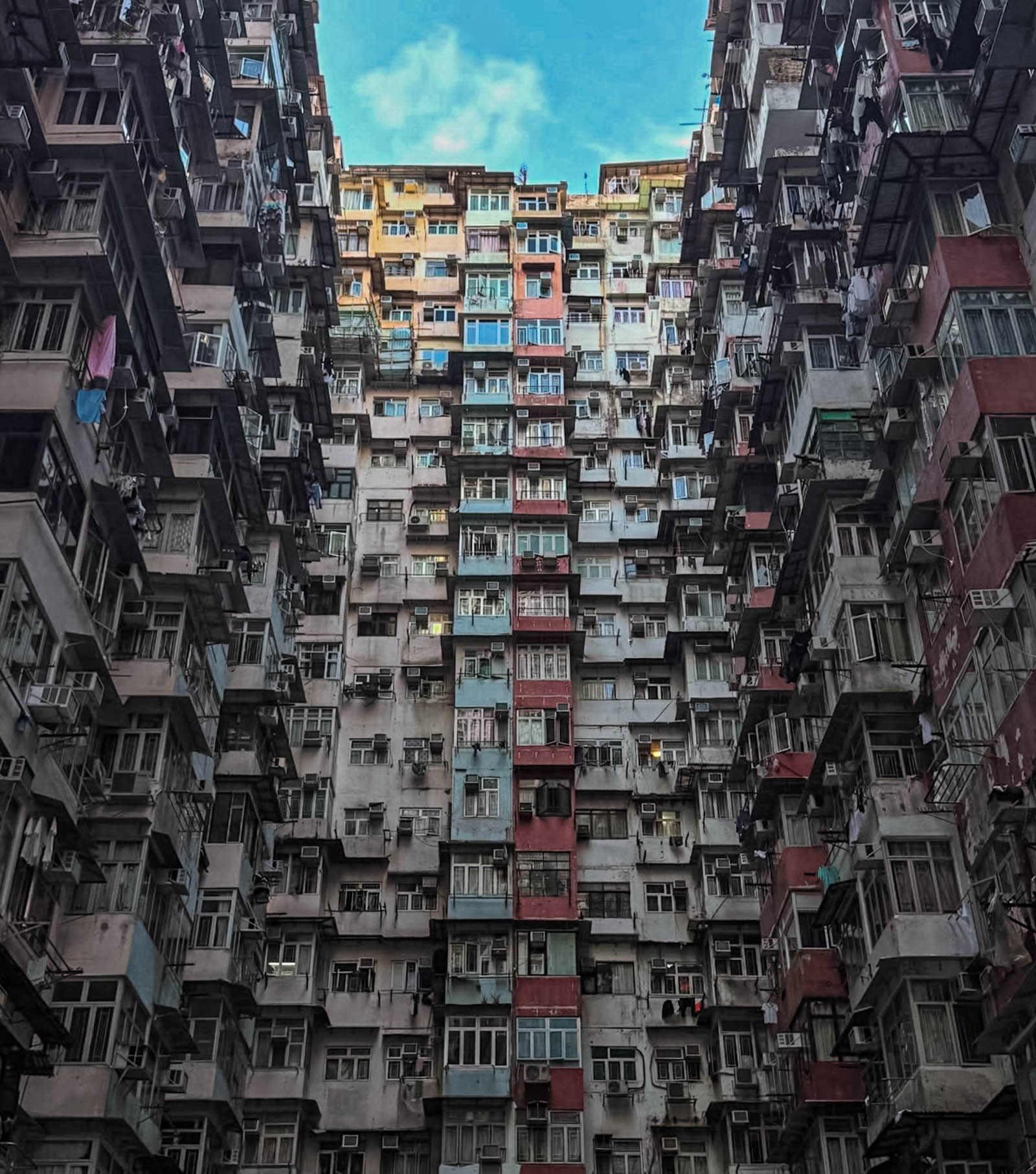 Looking up inside “Monster Building” residential complex – A vertigo-inducing view from the courtyard of Yick Cheong “Monster” Building: the camera gazes straight up at a narrow opening of sky framed by crowded, weathered apartment blocks on all four sides. Countless windows, laundry racks, and AC units form layers of a concrete canyon, epitomizing Hong Kong’s extreme housing density (Hong Kong travel, Monster Building, urban density, architecture).