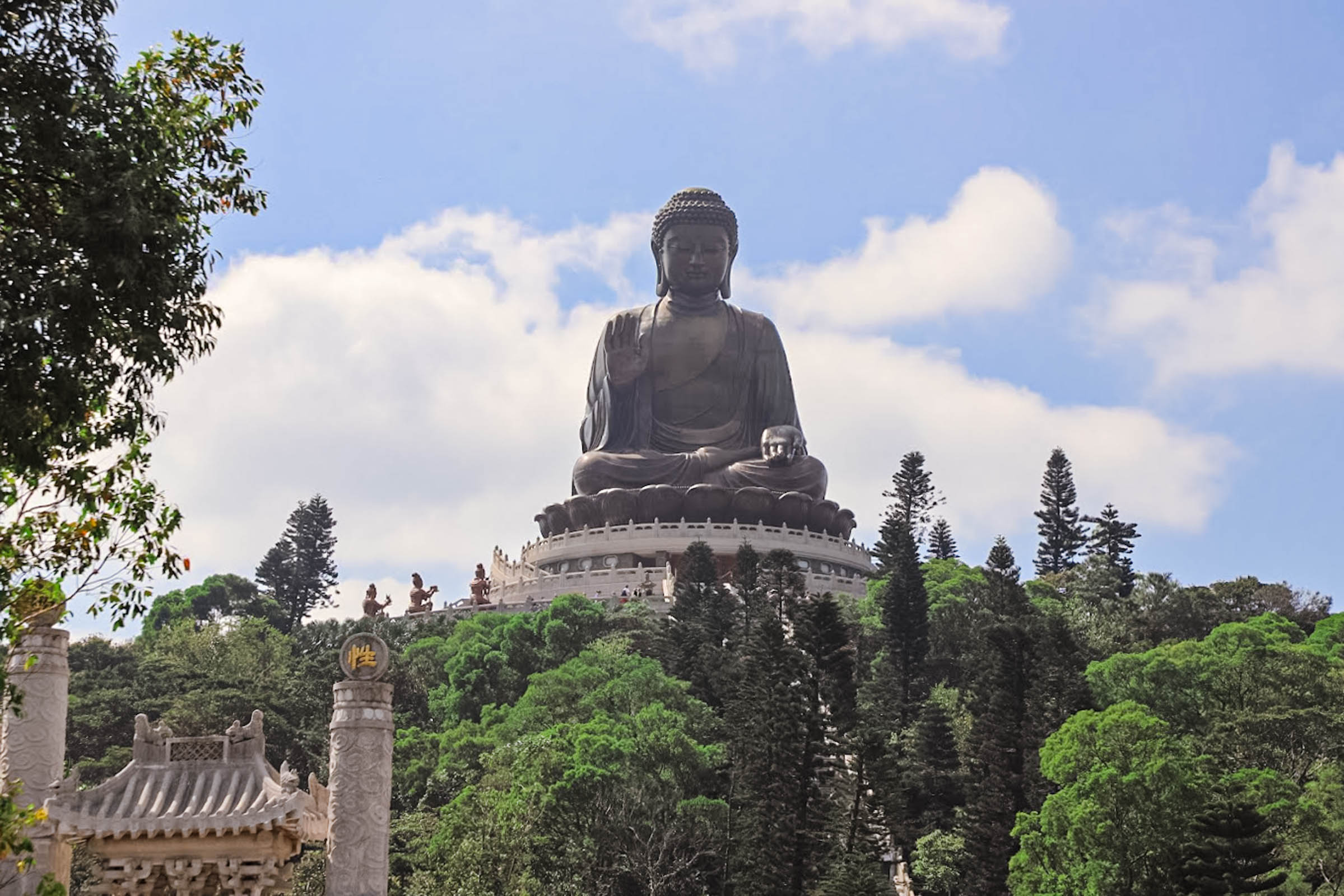 The Big Buddha statue on Lantau Island – The 34-meter bronze Tian Tan Buddha towers atop a hill at Ngong Ping, facing outward with a gentle smile and right hand raised in blessing. Tiny figures of visitors climb the 268 steps toward the statue’s lotus pedestal, conveying its immense scale and spiritual significance under a bright sky (Hong Kong travel, Big Buddha, Lantau Island, religious landmark).