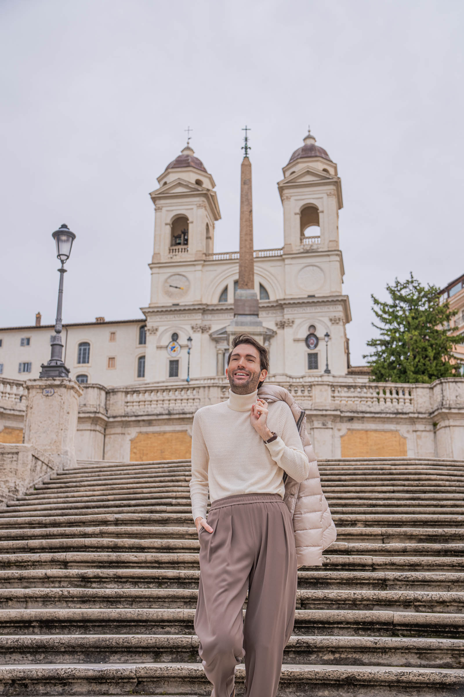 Marko Tadić, founder of Mr.M by Marko Tadić, standing on the Spanish Steps in Rome in front of Trinita dei Monti church with Egyptian obelisk, beige Herno quilted down jacket and Giorgio Armani trousers, Briston watch visible on wrist