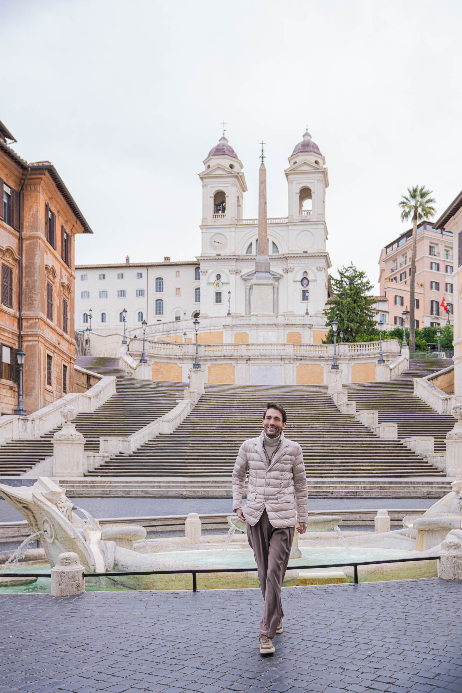 Marko Tadić, founder of Mr.M by Marko Tadić, full-length shot walking in front of the Spanish Steps in Rome, wearing a beige Herno quilted down jacket with sheen, Giorgio Armani cashmere turtleneck and wide-leg trousers with Fratelli Rossetti sneakers, Trinita dei Monti church in the background