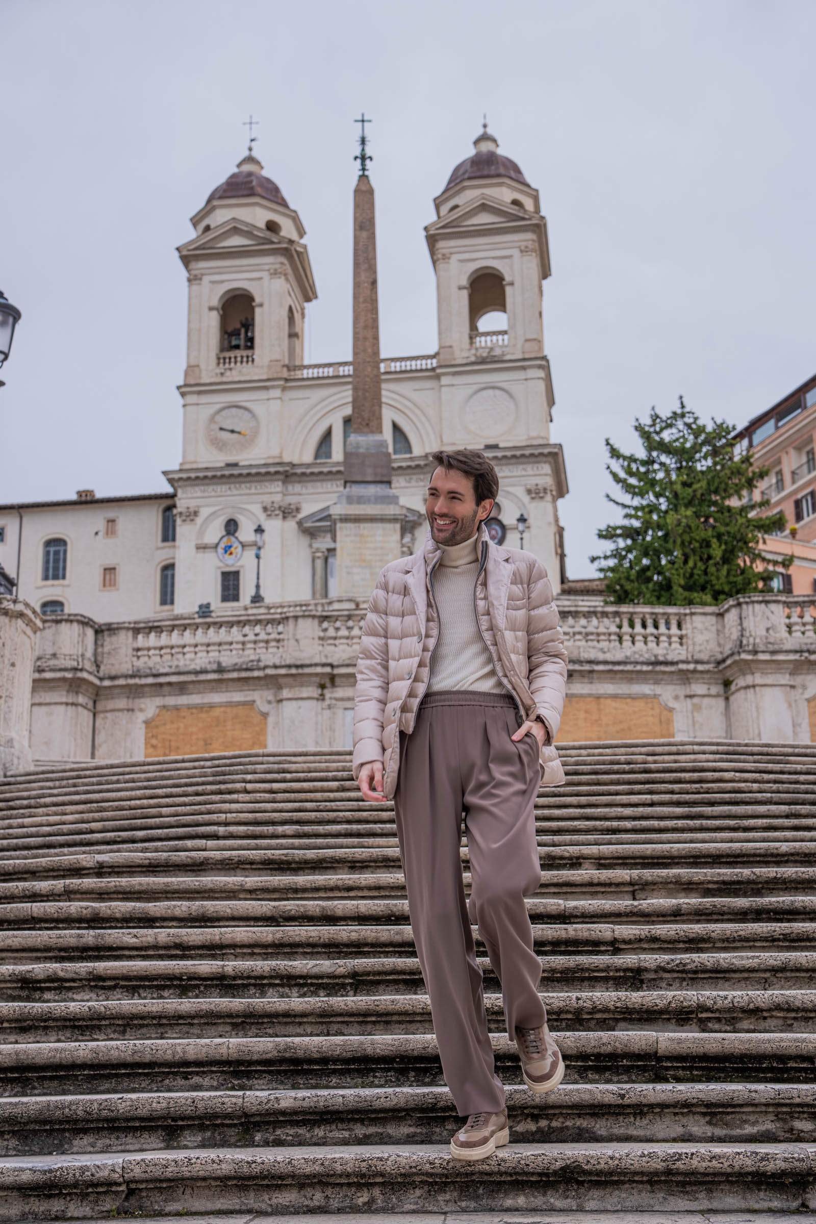 Marko Tadić, founder of Mr.M by Marko Tadić, standing on the Spanish Steps in Rome with Herno jacket draped over shoulder, Giorgio Armani cashmere turtleneck and wide-leg trousers, Briston watch on wrist, Trinita dei Monti church and obelisk in the background