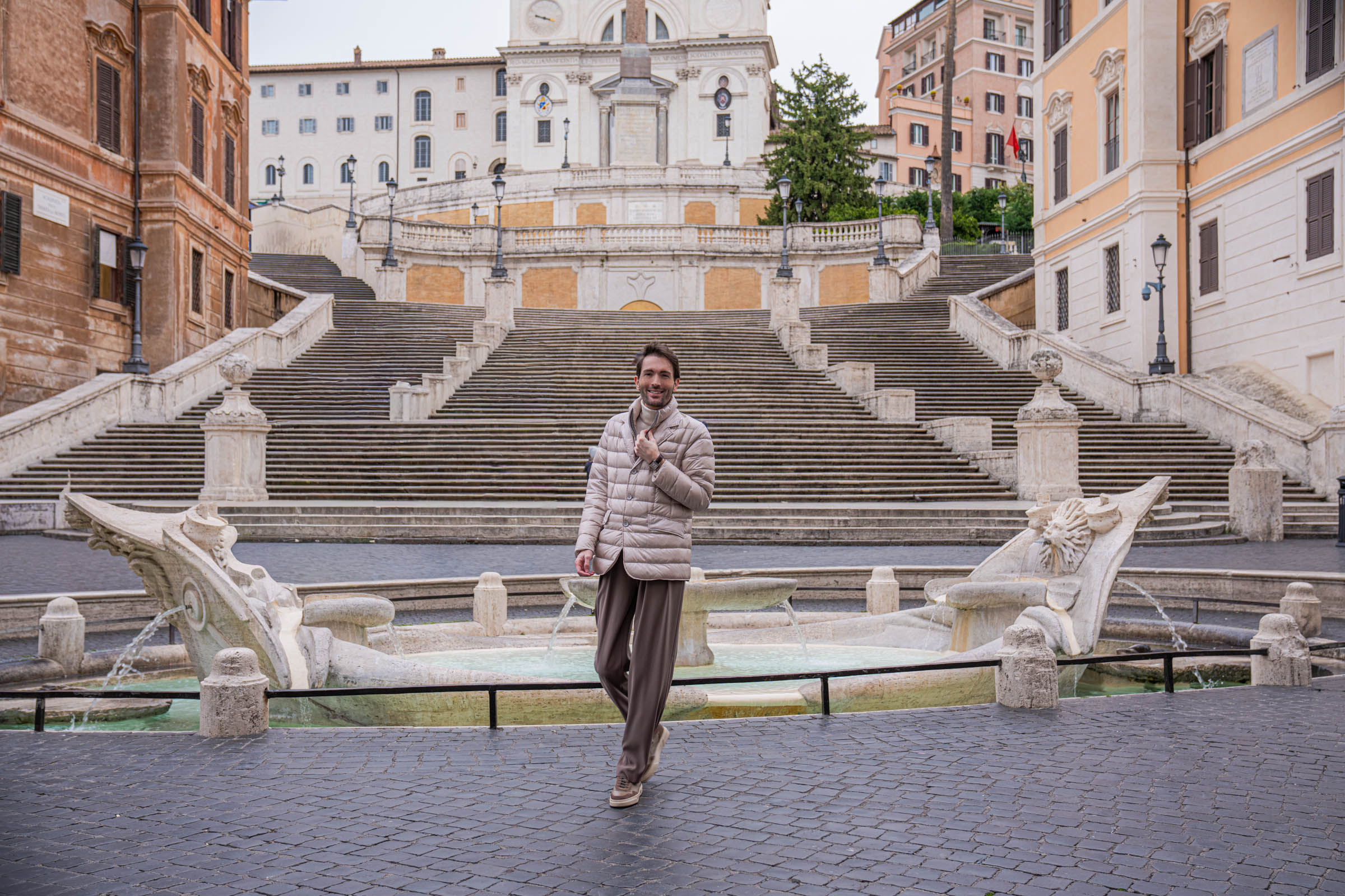 Marko Tadić, founder of Mr.M by Marko Tadić, walking past the Fontana della Barcaccia on Piazza di Spagna in Rome, beige Herno jacket open over Giorgio Armani cashmere turtleneck, Briston Streamliner Kennedy watch on wrist, Trinita dei Monti church behind