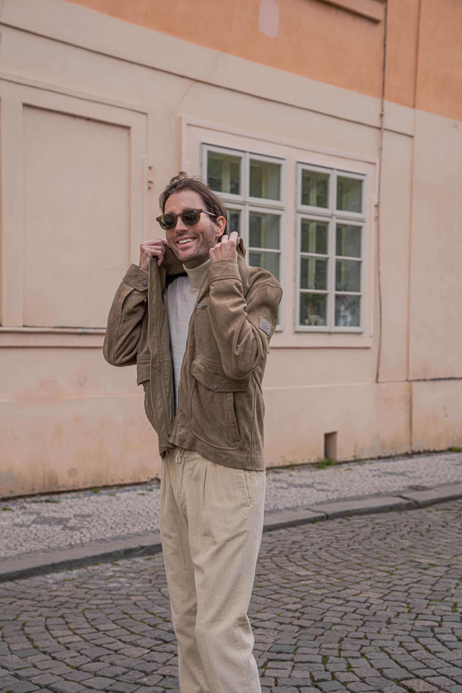 Marko Tadić, founder of Mr.M by Marko Tadić blog, side profile with popped collar in a Belstaff suede aviator jacket against a peach Baroque facade of Prague.