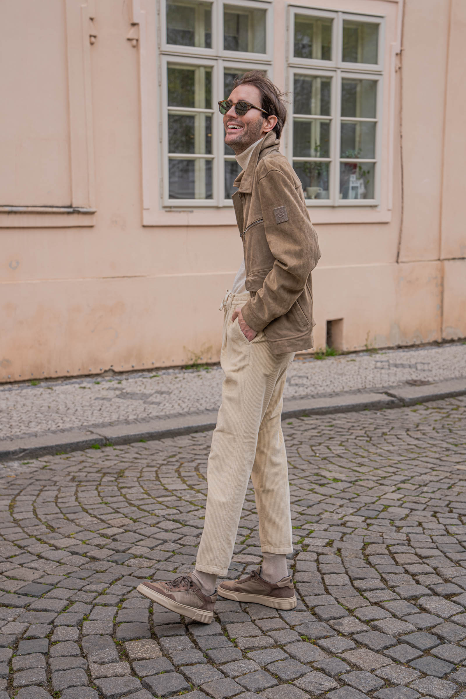 Marko Tadić, founder of Mr.M by Marko Tadić blog, smiling three-quarter portrait in a Belstaff suede aviator jacket against a weathered cream Prague facade.