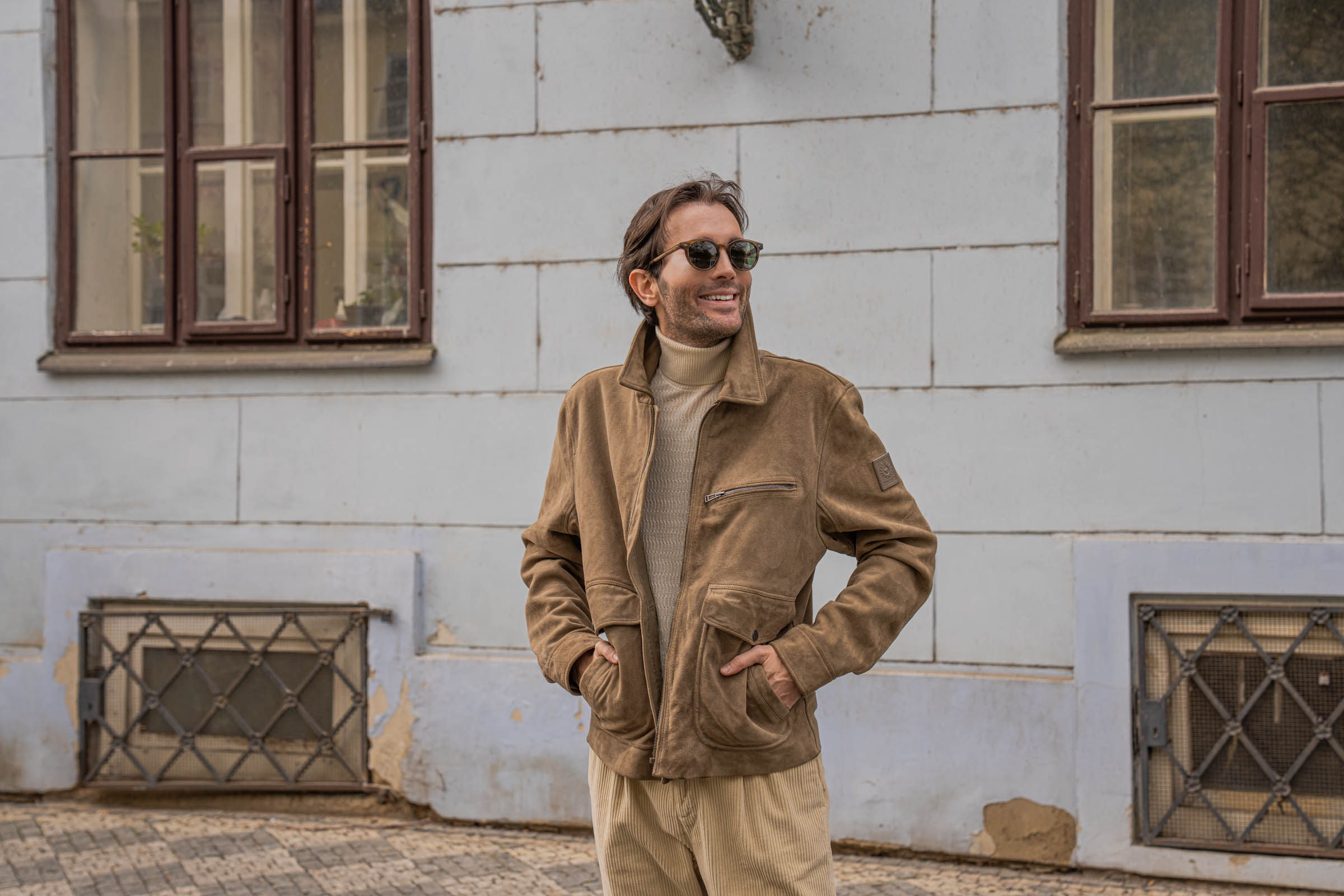 Marko Tadić, founder of Mr.M by Marko Tadić blog, standing pose adjusting tortoise sunglasses in a Belstaff suede jacket on the quiet streets of Malá Strana.