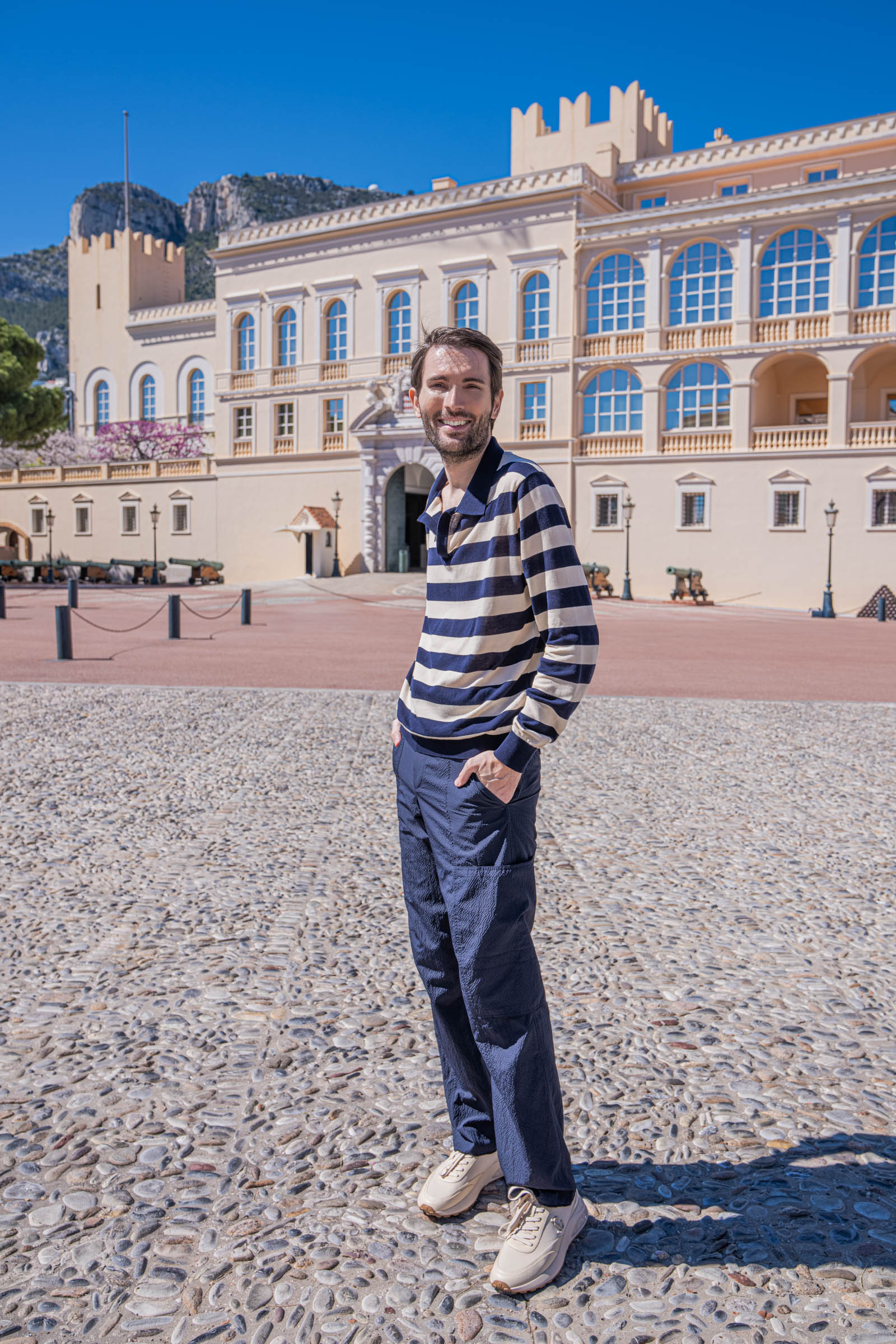 Marko Tadić, founder of the Mr.M blog, in a FERRAGAMO navy and cream striped silk knit polo shirt and navy cargo trousers standing on the pebble mosaic square in front of the Prince's Palace of Monaco