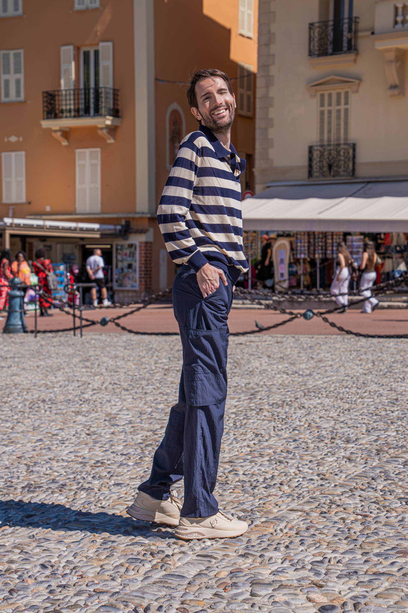 Marko Tadić, founder of the Mr.M blog, smiling over his shoulder in a FERRAGAMO striped long sleeve knit polo, photographed at Place du Palais in Monaco with the palace tower and blossoming spring trees behind him