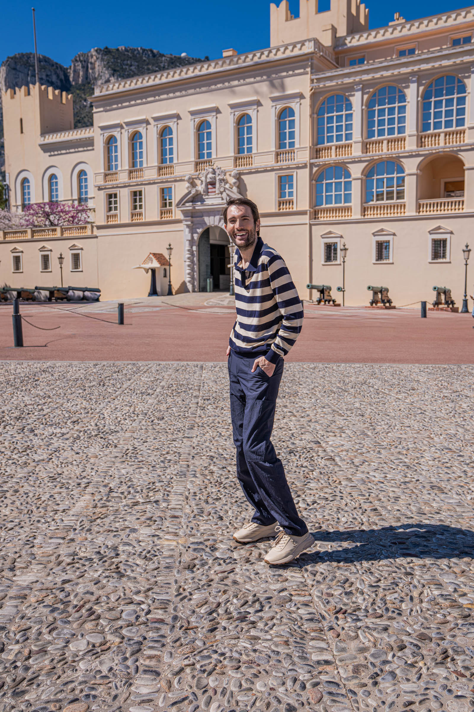 Marko Tadić, founder of the Mr.M blog, in an elegant casual look with FERRAGAMO striped silk polo and navy seersucker cargo pants, historic cannons and the cream facade of Monaco's royal palace behind him