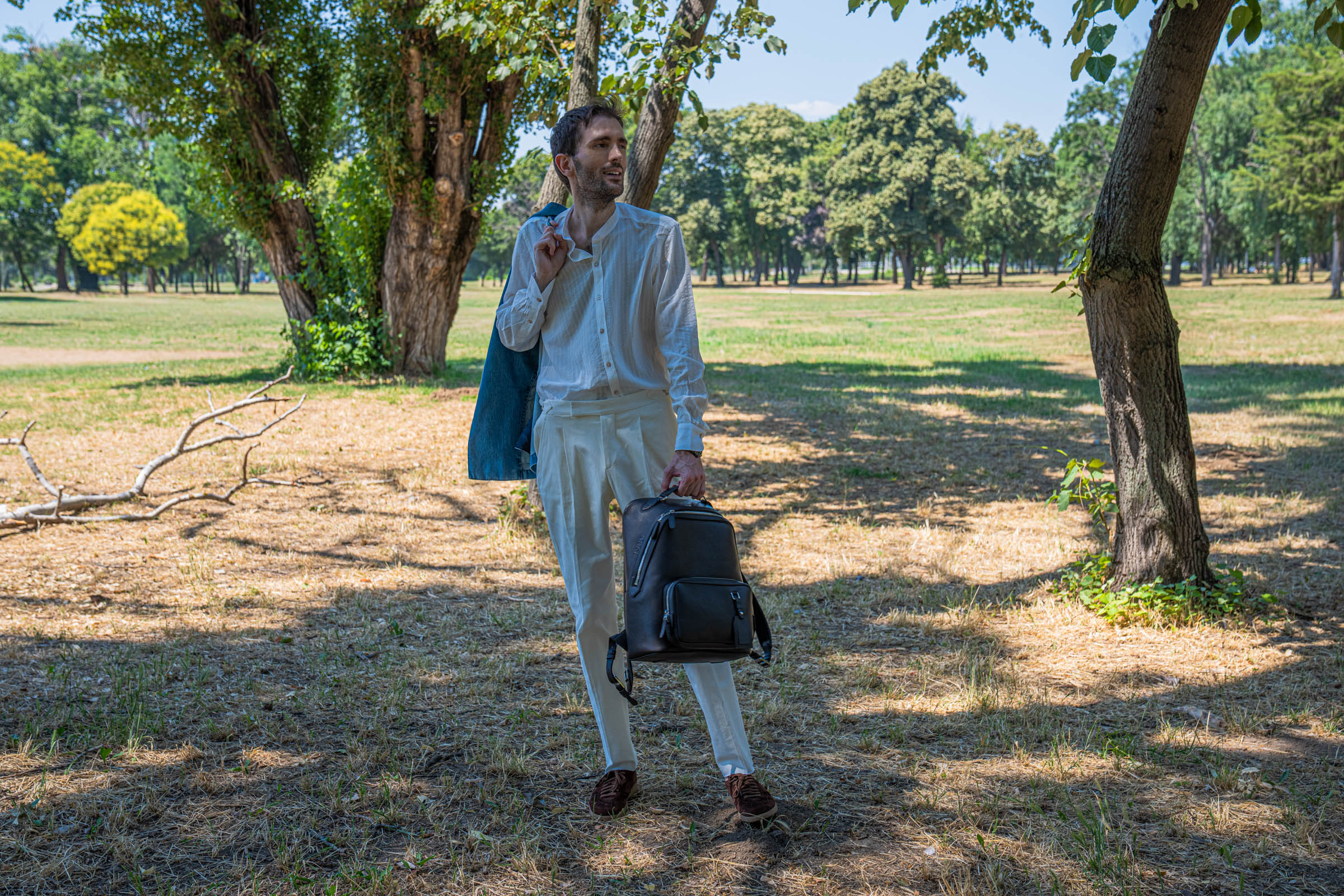 Fashion blogger Marko Tadić strolling through a city park with a grained leather 1900 backpack, denim blazer over his shoulder and a tonal cream outfit underneath