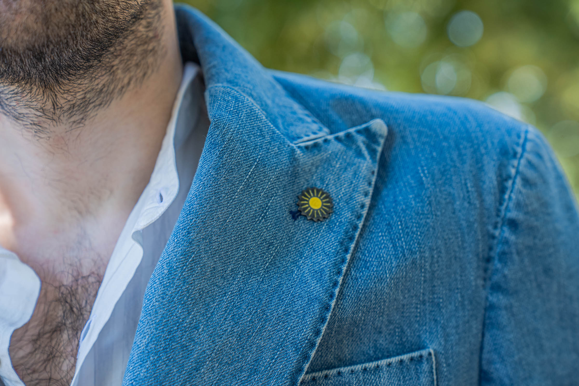 Close-up of the yellow sun pin on the peak lapel of Marko Tadić's light-wash Italian denim jacket, the signature detail of Giannetto Portofino