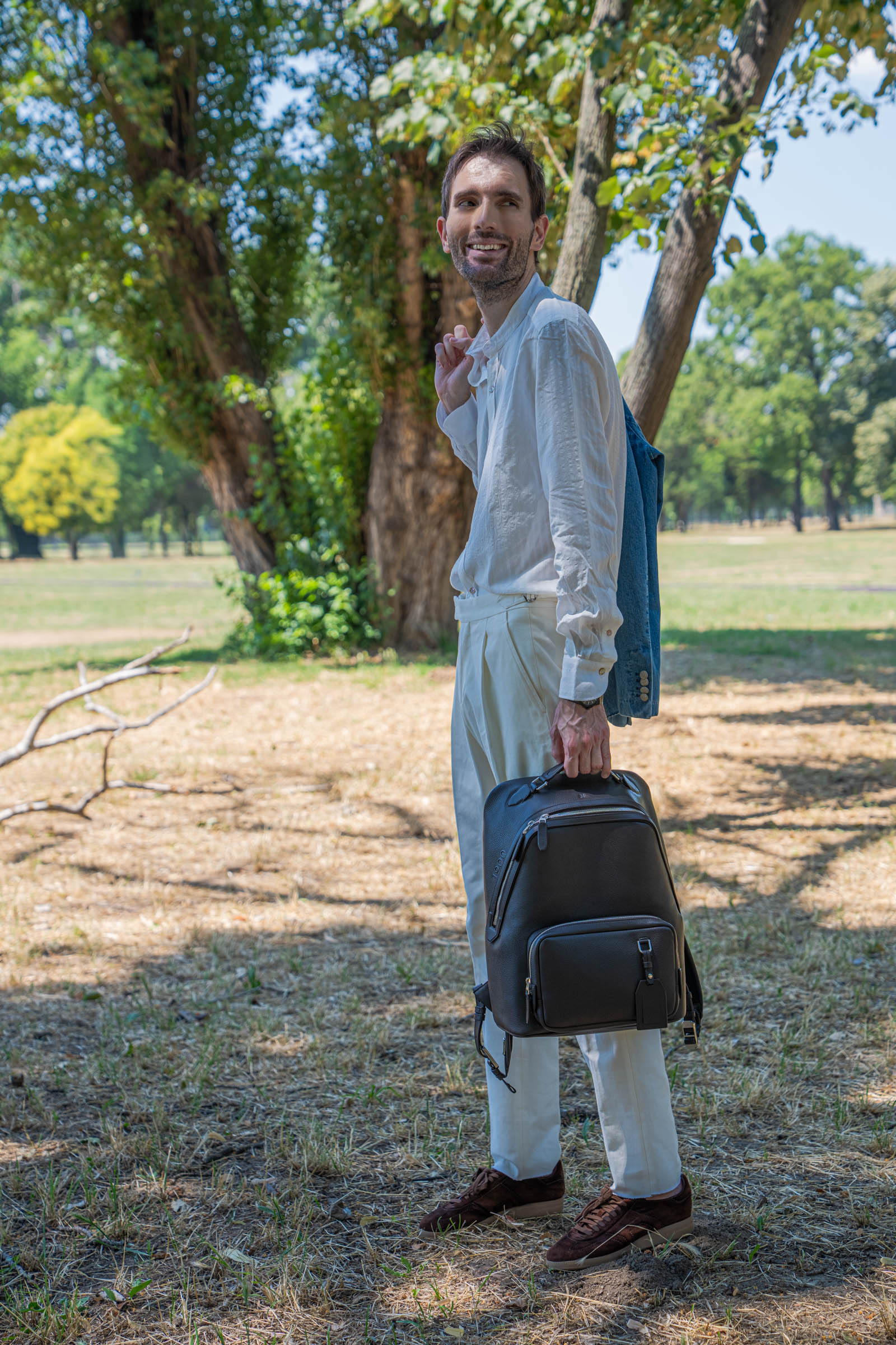 Smiling menswear blogger Marko Tadić carrying the 1900 Tempo Grande backpack by its top handle, wearing an ivory band collar shirt with a light-wash denim jacket draped over his shoulder