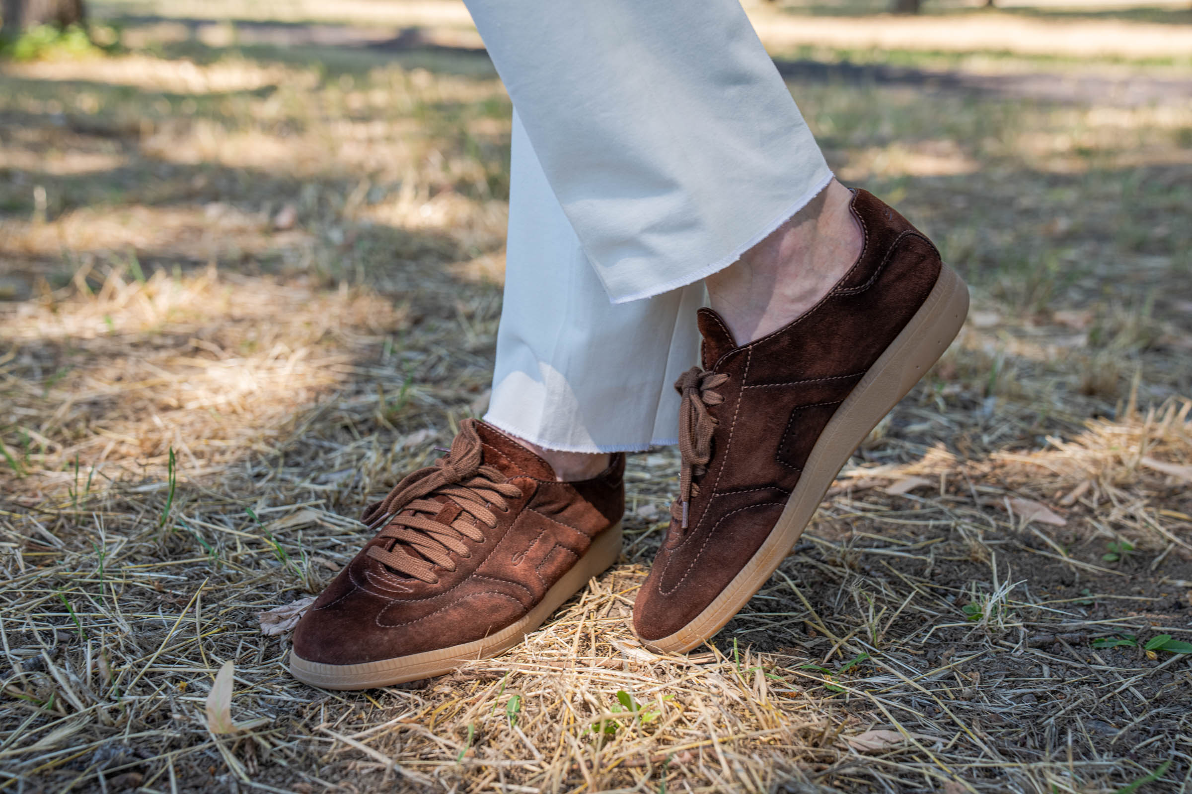 Close-up of Marko Tadić's Santoni DBS Oly sneakers in hand-gradated brown suede with natural gum rubber soles on dry summer grass