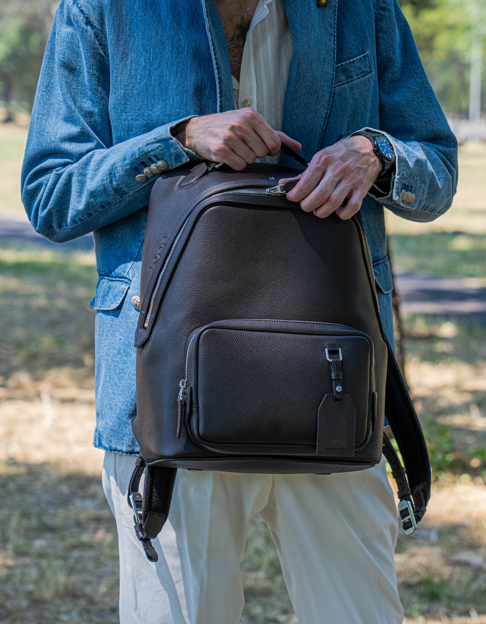 Marko Tadić in a denim blazer unzipping the front pocket of the dark brown 1900 Tempo Grande leather backpack held in front of him