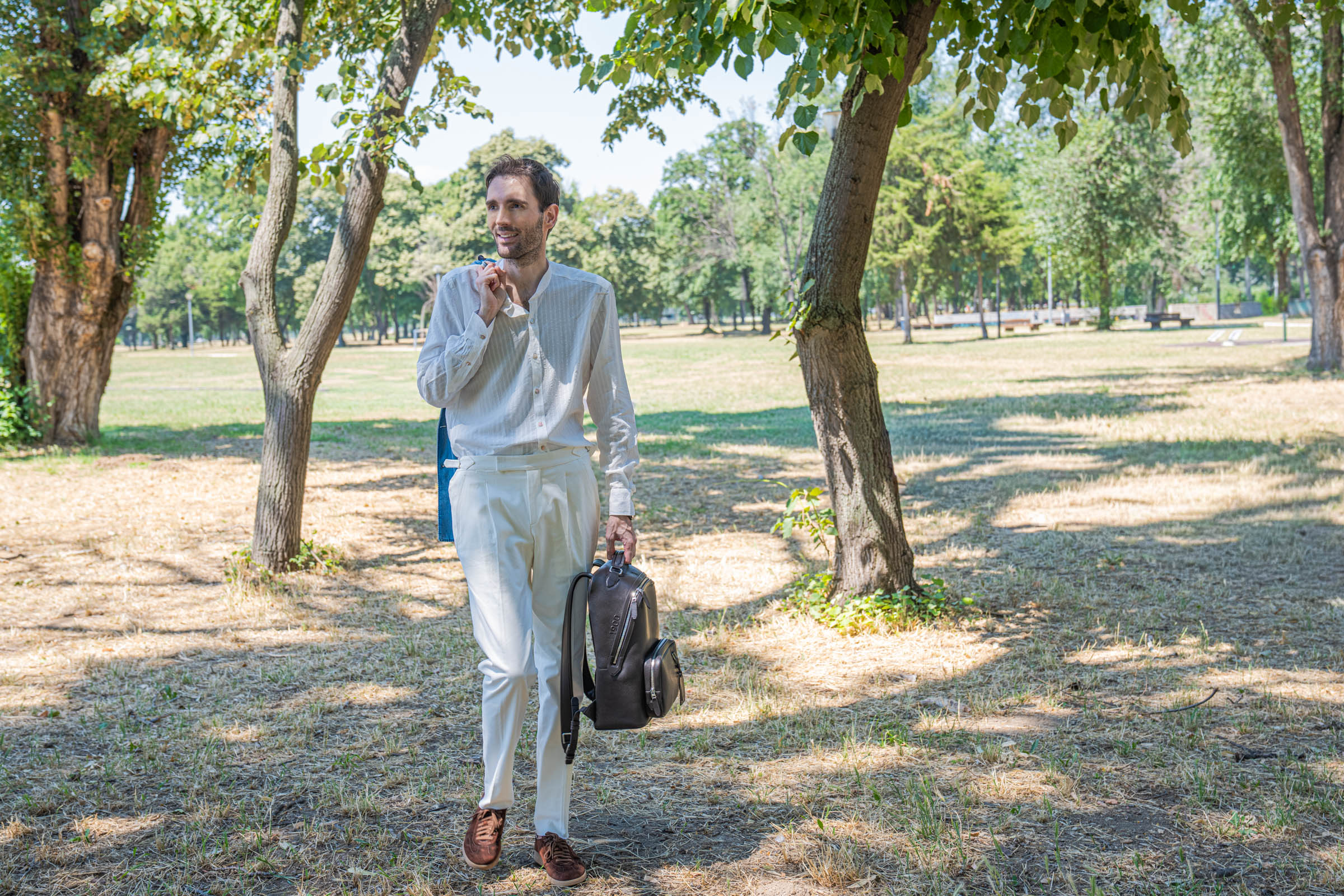 Marko Tadić of Mr.M walking through the park in a white lyocell shirt, cream pleated trousers and brown suede Santoni sneakers, dark brown leather backpack in hand