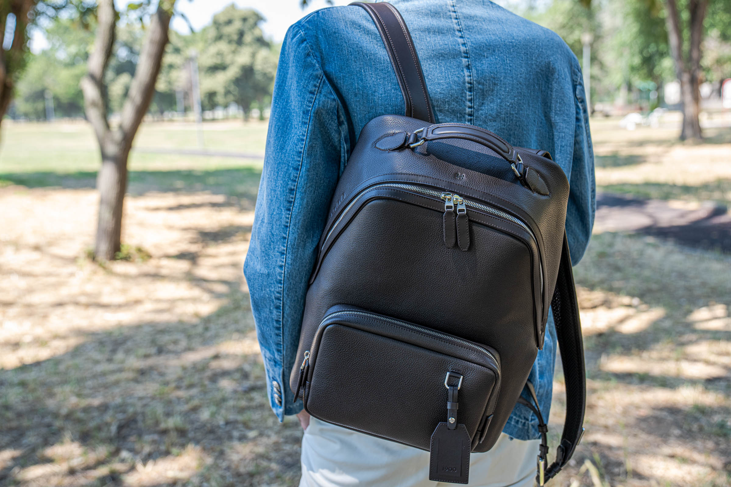Dark brown pebbled leather 1900 backpack worn over the shoulder of Marko Tadić's light-wash denim jacket, silver double zippers catching the sun