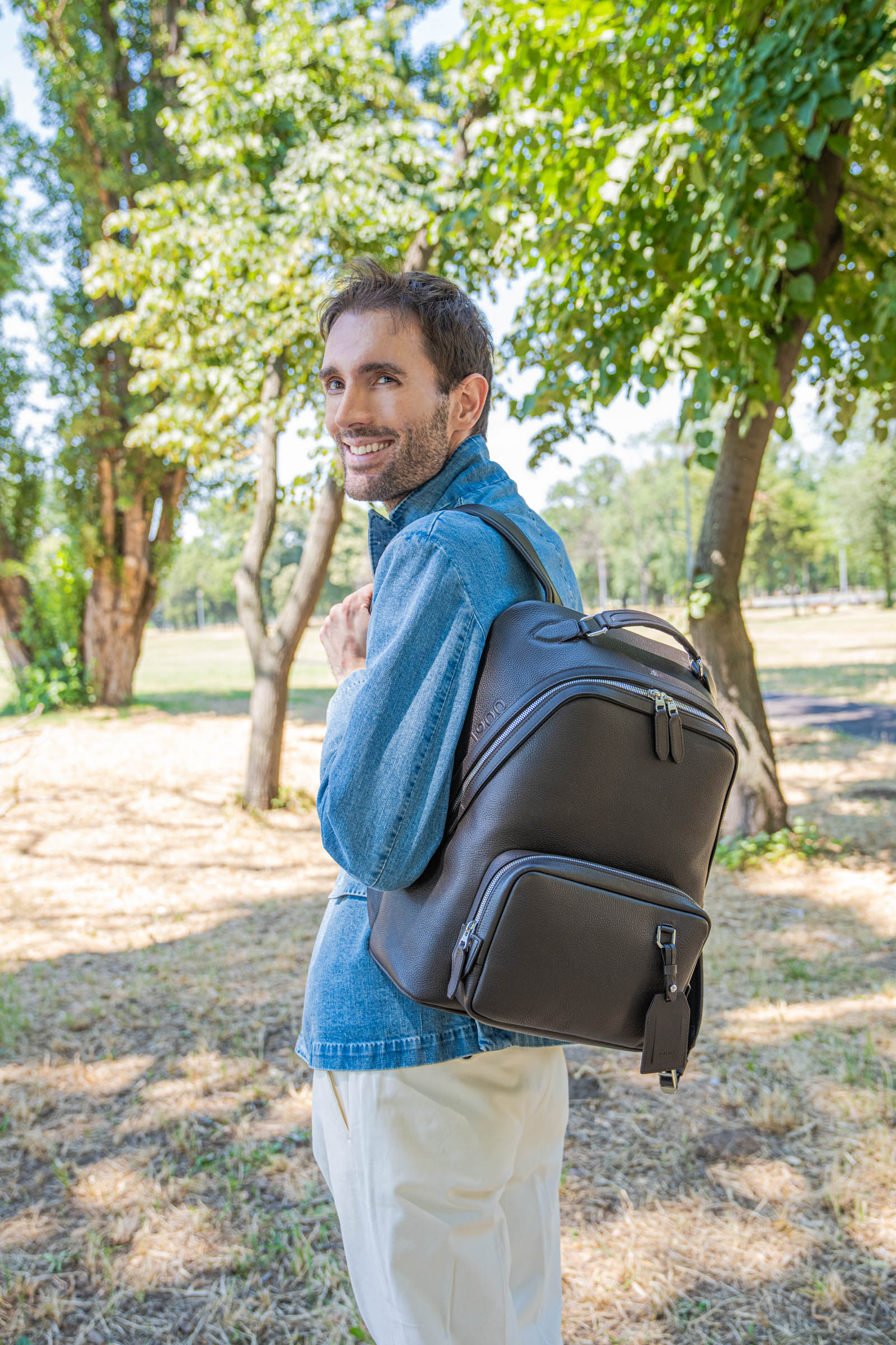 Side profile of a laughing Marko Tadić in a light blue denim jacket and off-white trousers strolling beneath summer trees