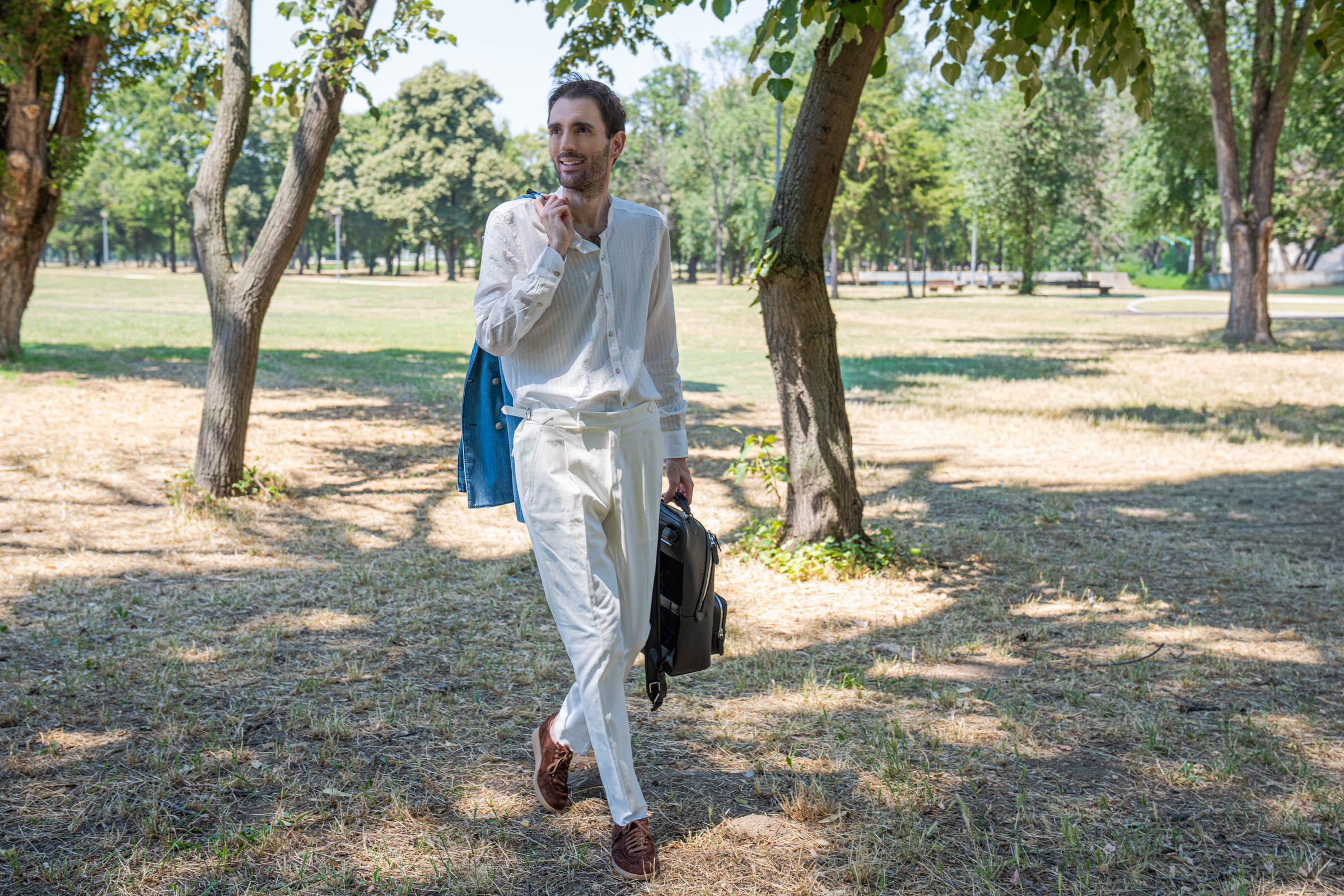 Marko Tadić in a white lyocell mandarin collar shirt and cream Cifonelli trousers holding a brown leather 1900 backpack under tall park trees, his Giannetto Portofino denim blazer slung over one shoulder