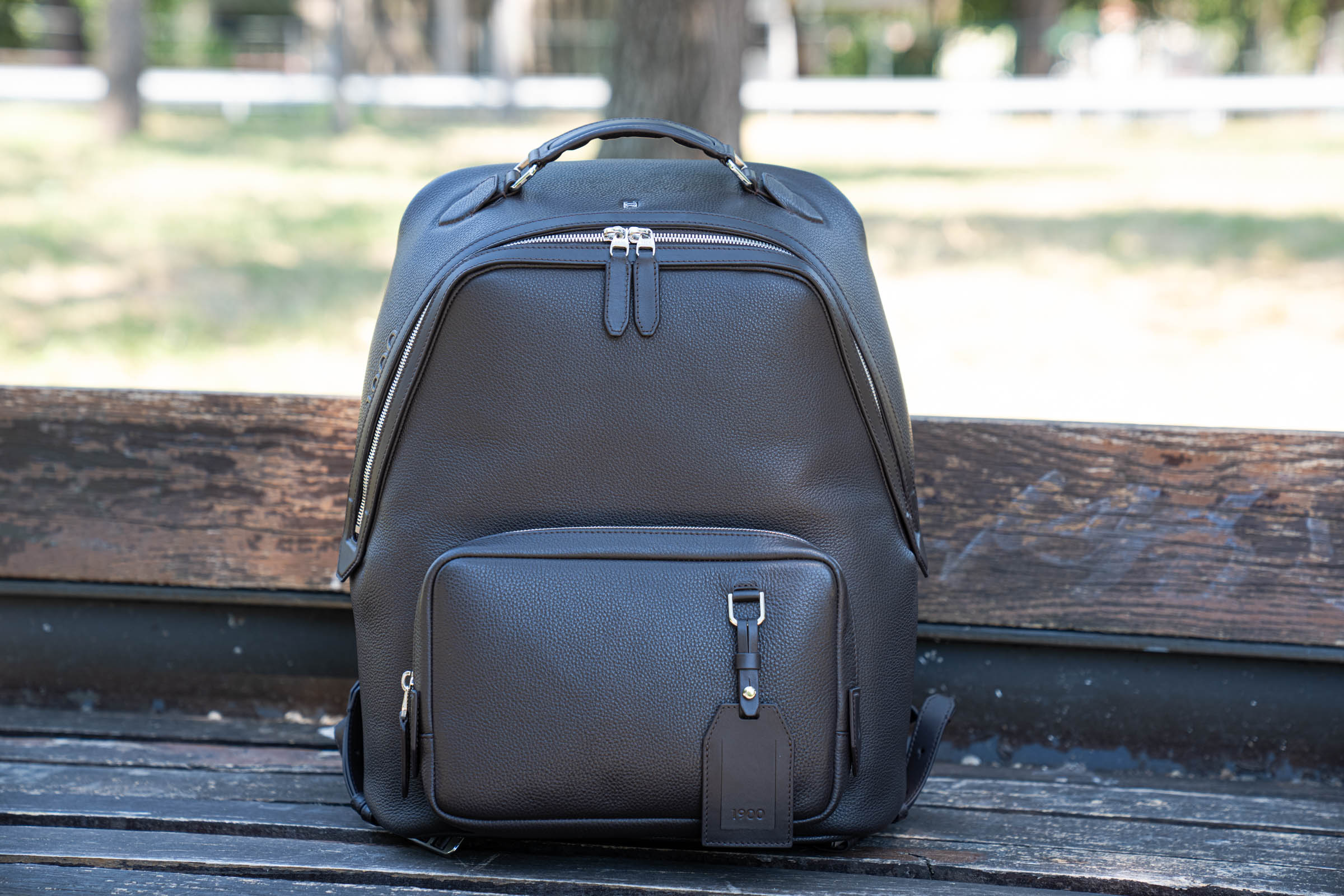 The 1900 Tempo Grande backpack in dark brown grained leather standing on a weathered wooden park bench, photographed from the front