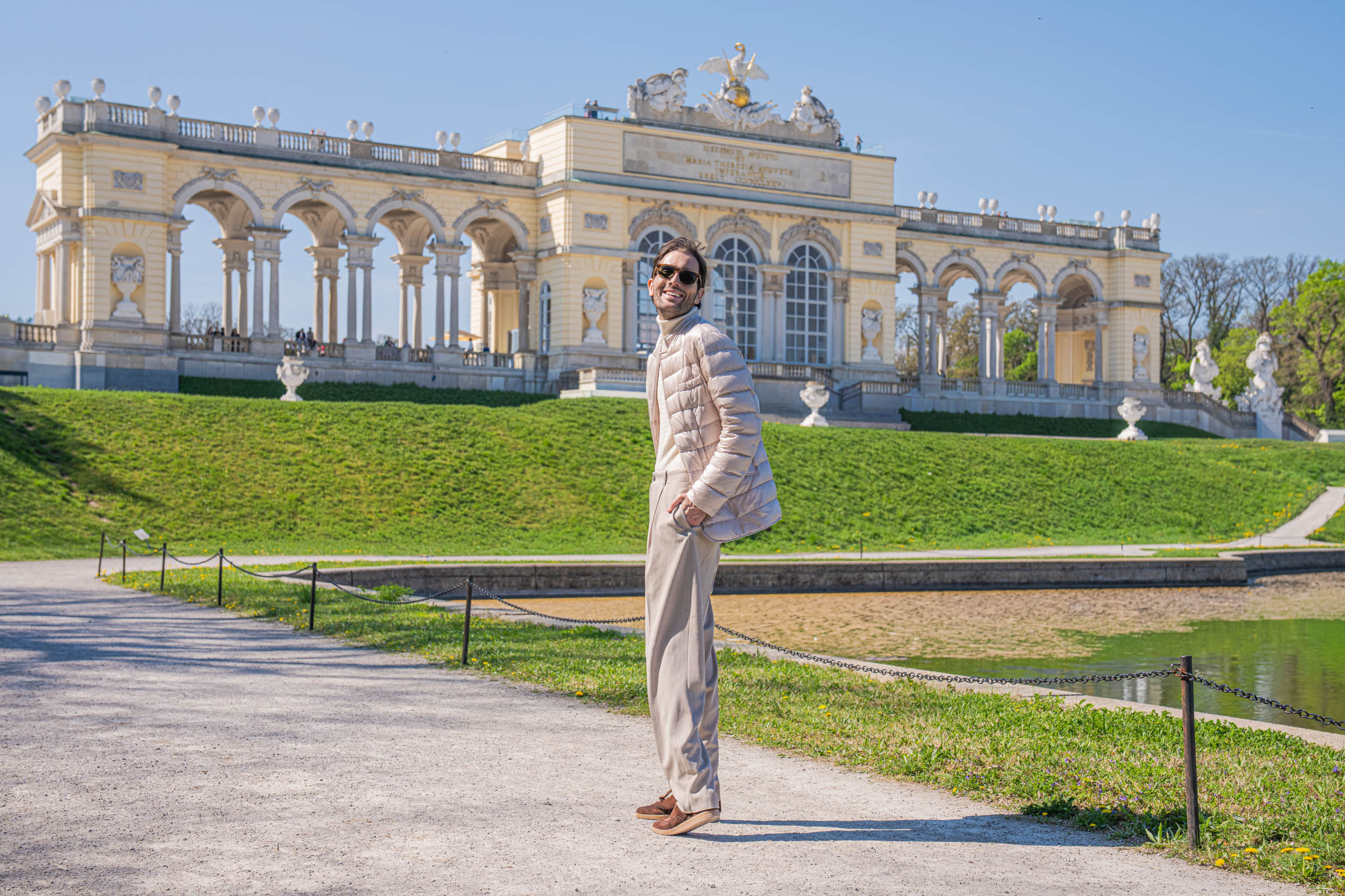Marko Tadić, founder of the Mr.M blog, in a playful side pose wearing Giorgio Armani style beige menswear below the Gloriette in Vienna